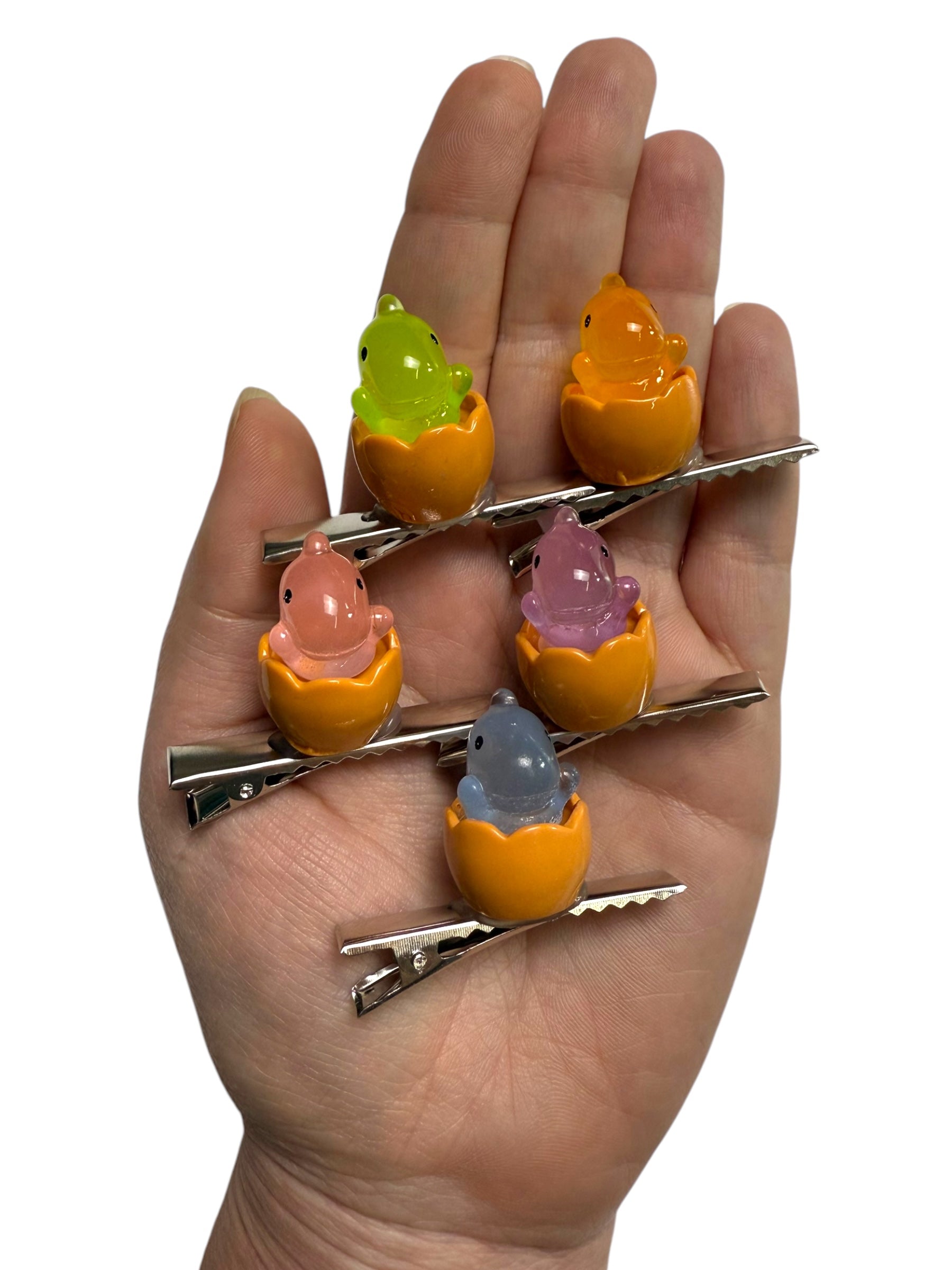 A hand holding five sprout hair clips, each featuring a small glowing dinosaur hatching from an orange egg.