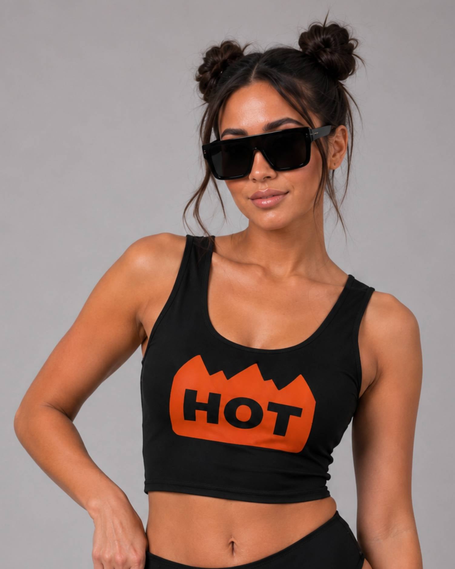 A woman wearing a black crop top tank with a bold orange graphic that says 'HOT' in black letters, styled with oversized black sunglasses against a solid gray studio background.