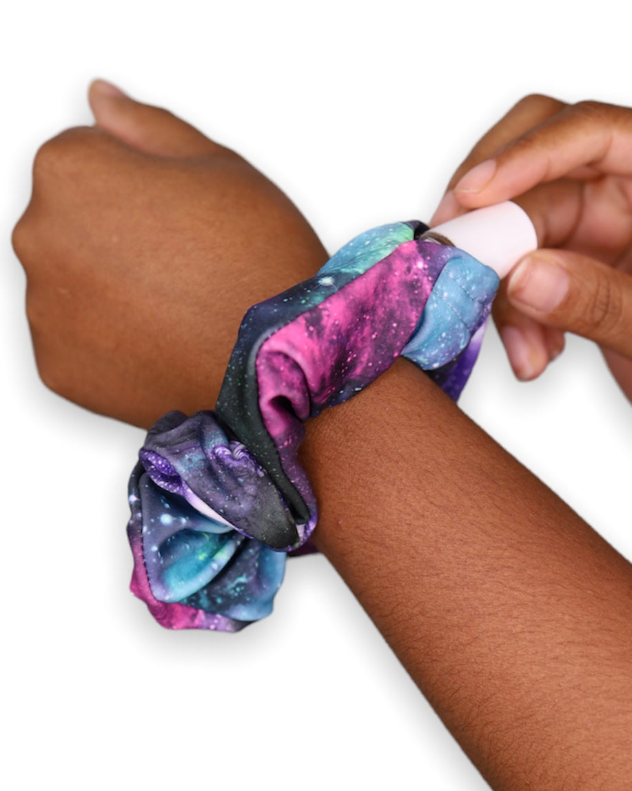 The Space Dust Stashie worn on the wrist, showcasing its deep space galaxy print and hidden Velcro® pocket storing lip balm.
