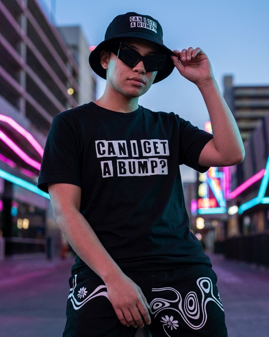 Person wearing a black t-shirt with text and black cap in an urban setting with neon lights.